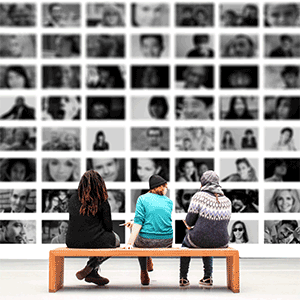 Picture of three people sitting on a bench facing a wall of portraits.
